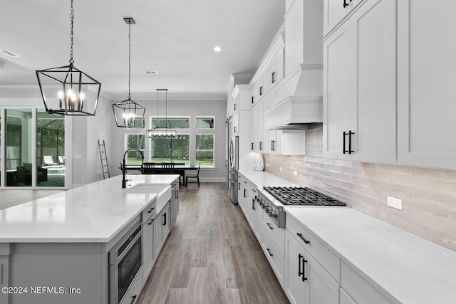 kitchen featuring white cabinetry, hanging light fixtures, stainless steel appliances, an island with sink, and custom exhaust hood