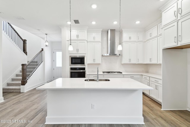 kitchen featuring stainless steel appliances, a sink, light countertops, wall chimney range hood, and an island with sink