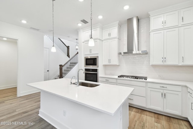 kitchen featuring an island with sink, wall chimney exhaust hood, light countertops, pendant lighting, and a sink