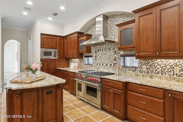 kitchen featuring brown cabinetry, light stone counters, a center island, stainless steel appliances, and wall chimney range hood