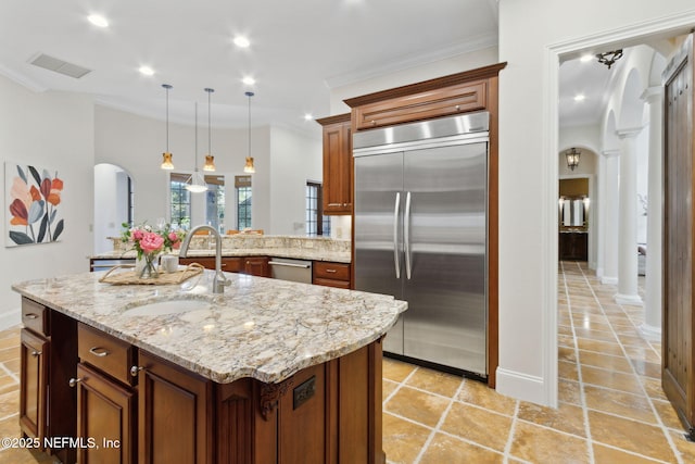 kitchen with arched walkways, a kitchen island with sink, a sink, appliances with stainless steel finishes, and brown cabinets