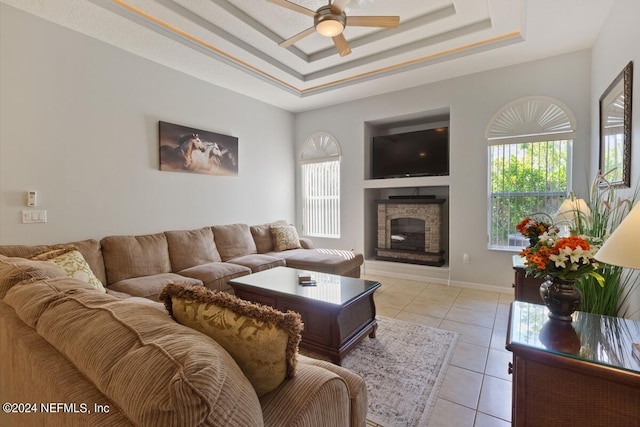 tiled living room featuring a raised ceiling, ceiling fan, and a stone fireplace