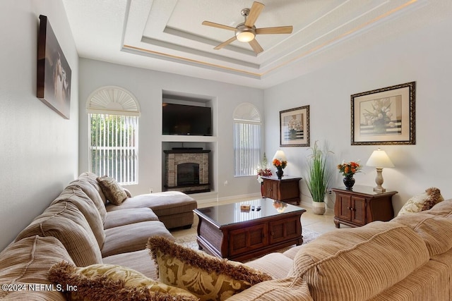 tiled living room featuring a tray ceiling, a stone fireplace, and ceiling fan