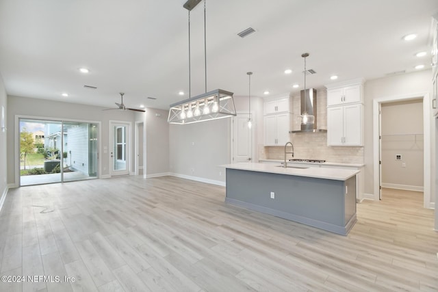 kitchen featuring light wood-type flooring, wall chimney exhaust hood, a kitchen island with sink, white cabinets, and hanging light fixtures