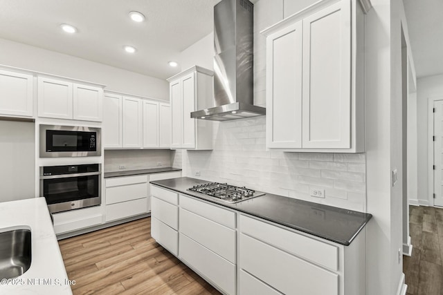 kitchen featuring stainless steel appliances, backsplash, light wood-style flooring, white cabinetry, and wall chimney range hood