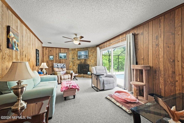 carpeted living room with wood walls, ceiling fan, crown molding, a stone fireplace, and a textured ceiling