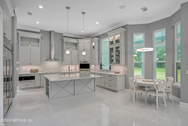 kitchen with a kitchen island with sink, wall chimney range hood, crown molding, decorative light fixtures, and light stone counters