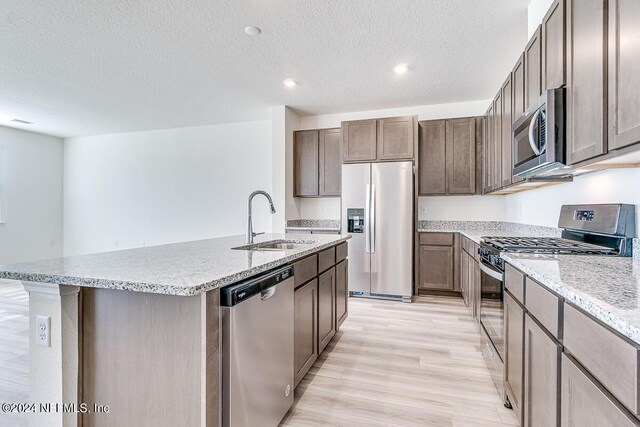 kitchen with appliances with stainless steel finishes, a kitchen island with sink, dark brown cabinets, light wood-type flooring, and a sink