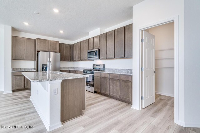 kitchen with dark brown cabinetry, appliances with stainless steel finishes, light stone countertops, a kitchen island with sink, and light wood-type flooring