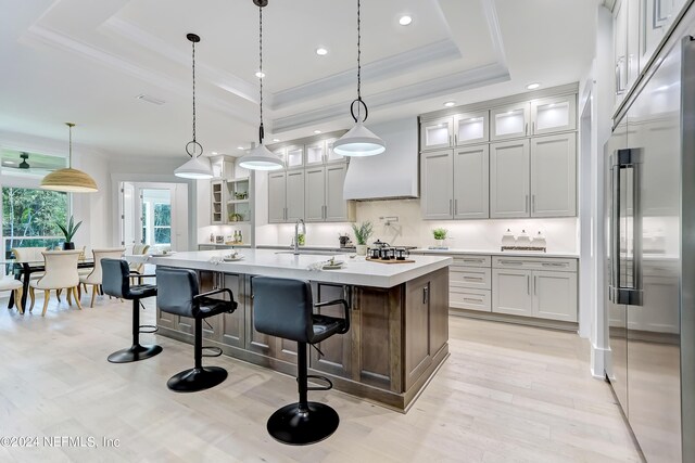 kitchen with a tray ceiling, high quality fridge, a center island with sink, pendant lighting, and premium range hood