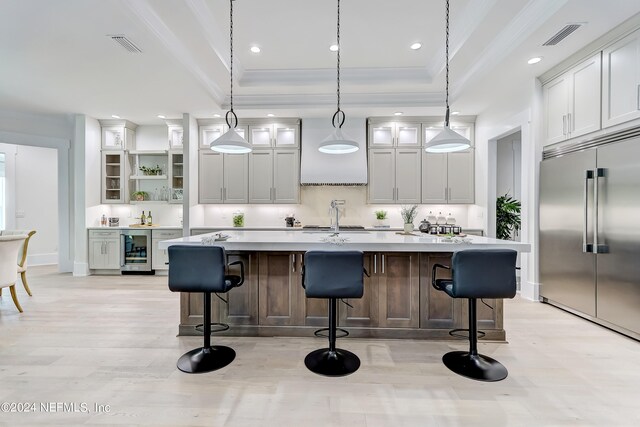 kitchen featuring stainless steel built in fridge, light hardwood / wood-style flooring, a kitchen island with sink, pendant lighting, and a raised ceiling