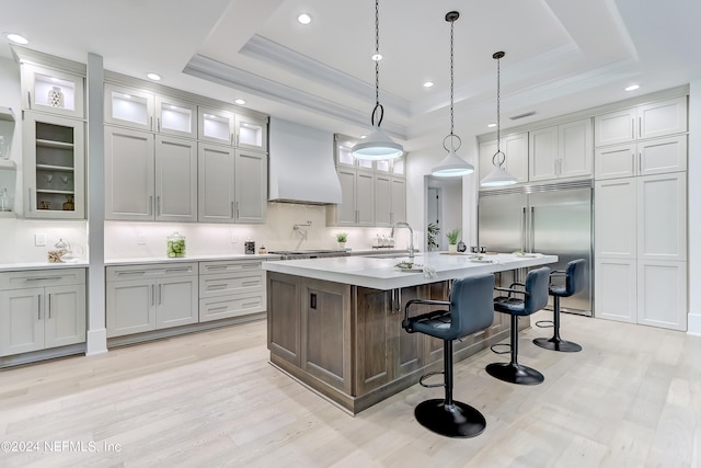 kitchen with a tray ceiling, light hardwood / wood-style flooring, custom exhaust hood, a center island with sink, and pendant lighting