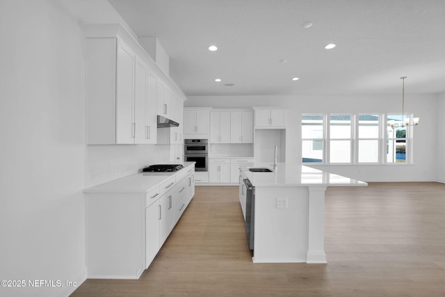 kitchen with white cabinets, appliances with stainless steel finishes, an island with sink, sink, and a notable chandelier