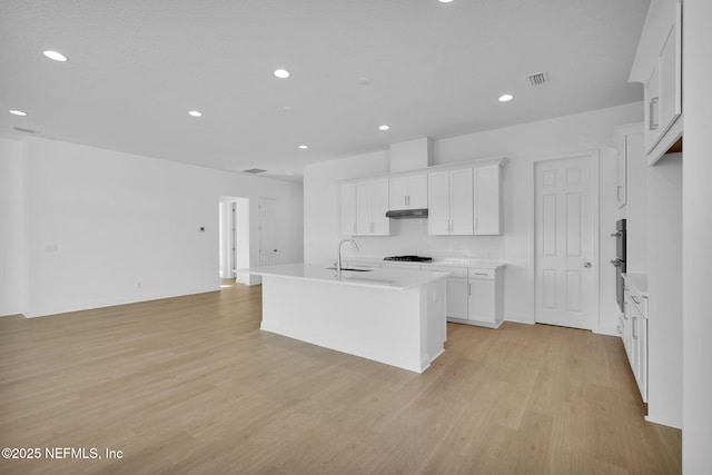 kitchen featuring white cabinets, sink, backsplash, a kitchen island with sink, and light wood-type flooring