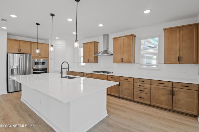 kitchen featuring appliances with stainless steel finishes, light wood-type flooring, sink, wall chimney range hood, and a center island with sink