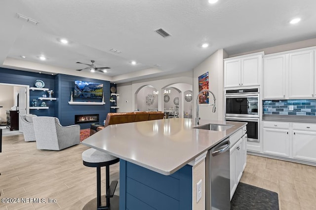 kitchen with a large fireplace, stainless steel appliances, ceiling fan, a center island with sink, and white cabinets