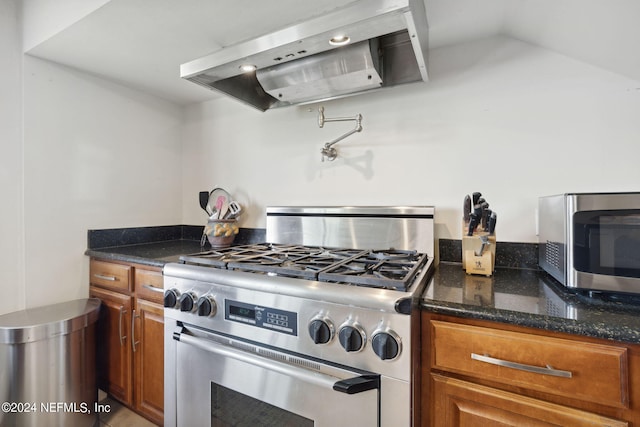 kitchen with stainless steel appliances, vaulted ceiling, range hood, brown cabinets, and dark stone countertops