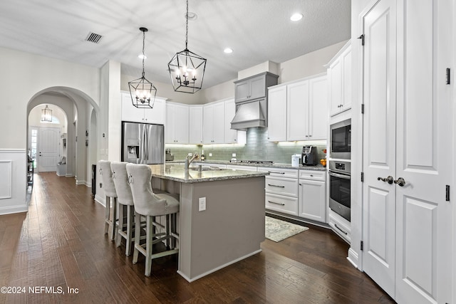 kitchen featuring custom exhaust hood, stainless steel appliances, an island with sink, backsplash, and dark wood-type flooring