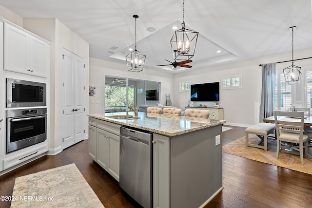 kitchen with appliances with stainless steel finishes, ceiling fan with notable chandelier, an island with sink, and a tray ceiling