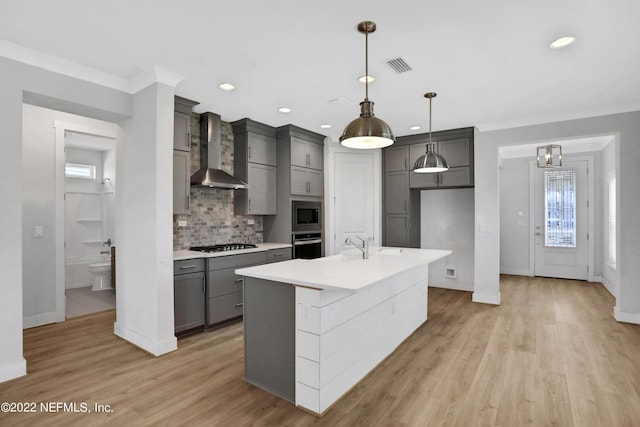 kitchen with decorative backsplash, hanging light fixtures, a kitchen island with sink, appliances with stainless steel finishes, and wall chimney exhaust hood