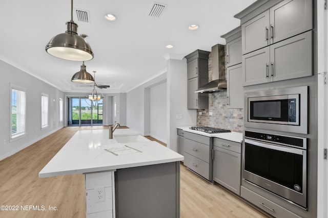 kitchen with an island with sink, appliances with stainless steel finishes, gray cabinetry, ornamental molding, and wall chimney exhaust hood