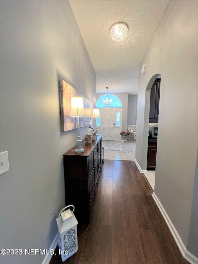 hall featuring a textured ceiling, arched walkways, baseboards, dark wood finished floors, and an inviting chandelier