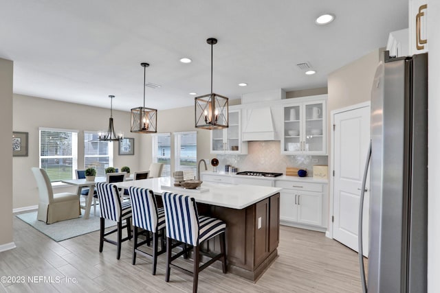 kitchen with white cabinetry, an island with sink, backsplash, decorative light fixtures, and stainless steel refrigerator