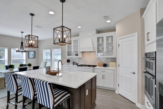 kitchen featuring premium range hood, white cabinetry, a kitchen island with sink, stainless steel gas cooktop, and hanging light fixtures