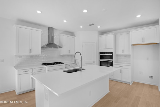 kitchen with white cabinetry, wall chimney exhaust hood, a center island with sink, and black appliances