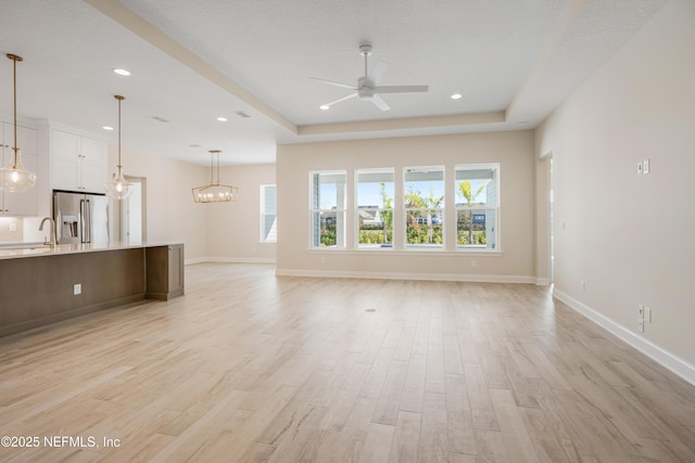 unfurnished living room featuring light wood finished floors, recessed lighting, a raised ceiling, baseboards, and ceiling fan with notable chandelier