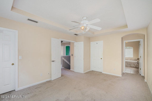 unfurnished bedroom with a tray ceiling, light colored carpet, ensuite bath, and ceiling fan