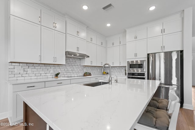 kitchen with appliances with stainless steel finishes, backsplash, light wood-type flooring, light stone countertops, and an island with sink