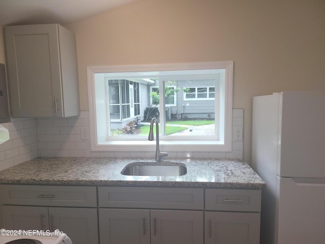 kitchen featuring decorative backsplash, freestanding refrigerator, light stone countertops, white cabinetry, and a sink