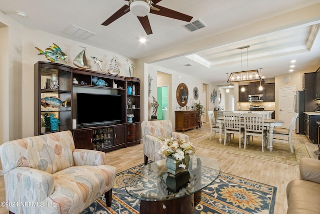 living room with ceiling fan with notable chandelier, a raised ceiling, and light wood-type flooring