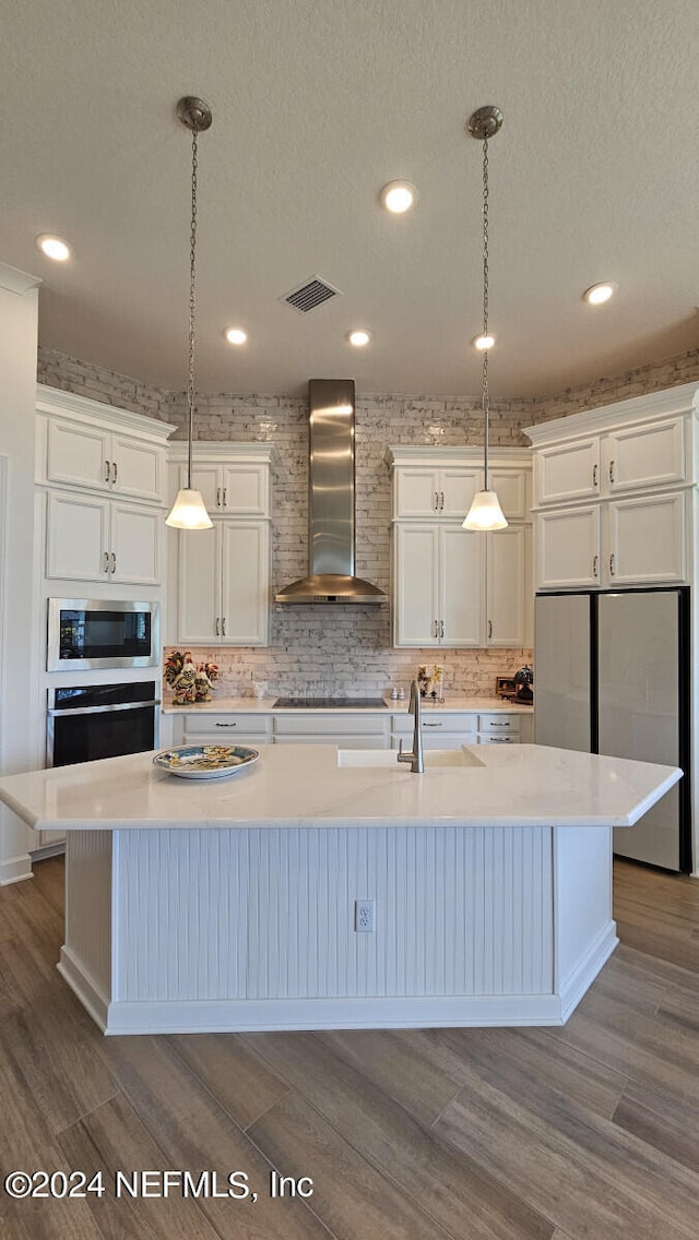 kitchen with decorative light fixtures, wall chimney range hood, a kitchen island with sink, and appliances with stainless steel finishes