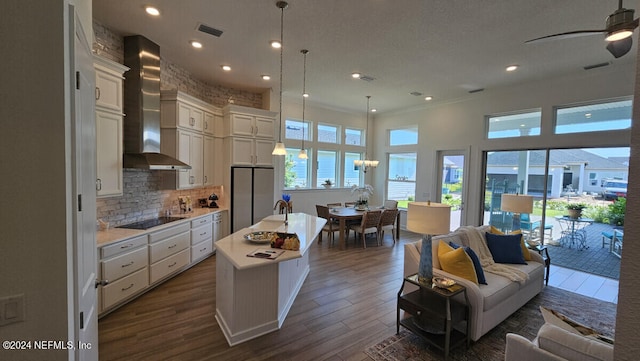 kitchen with hanging light fixtures, a center island with sink, wall chimney range hood, dark hardwood / wood-style flooring, and ceiling fan