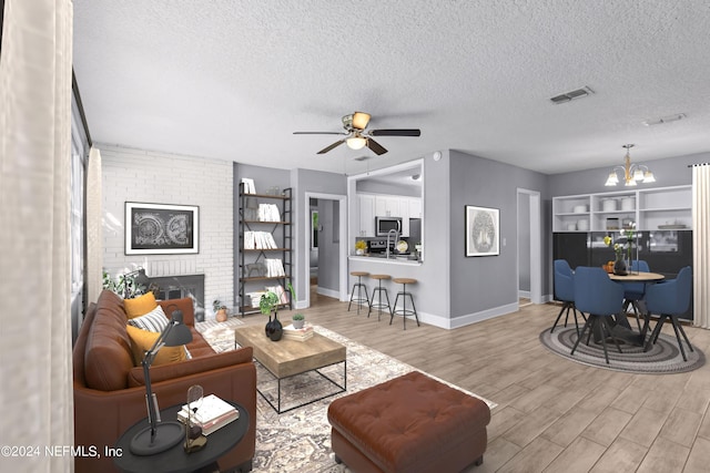 living room featuring a brick fireplace, ceiling fan with notable chandelier, a textured ceiling, and light hardwood / wood-style floors