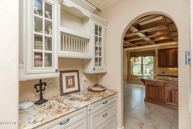 kitchen with arched walkways, coffered ceiling, glass insert cabinets, light stone counters, and crown molding