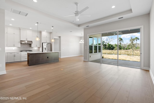 unfurnished living room with ceiling fan with notable chandelier, a tray ceiling, and light hardwood / wood-style floors