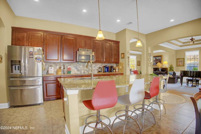 kitchen featuring tasteful backsplash, light stone counters, a kitchen island with sink, ceiling fan, and appliances with stainless steel finishes