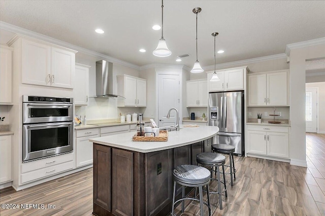 kitchen with appliances with stainless steel finishes, white cabinetry, wall chimney range hood, hanging light fixtures, and a center island with sink