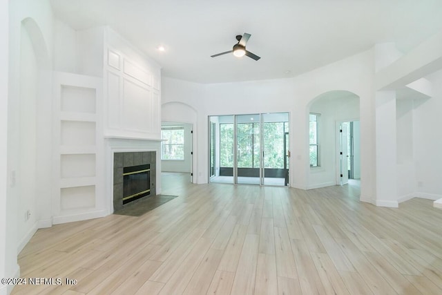 unfurnished living room featuring a tiled fireplace, light hardwood / wood-style flooring, ceiling fan, and built in shelves