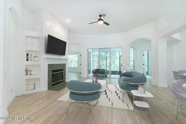 living room with built in shelves, ceiling fan, a tile fireplace, and light hardwood / wood-style flooring