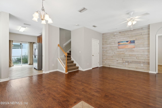 unfurnished living room with dark hardwood / wood-style floors, ceiling fan with notable chandelier, and wood walls