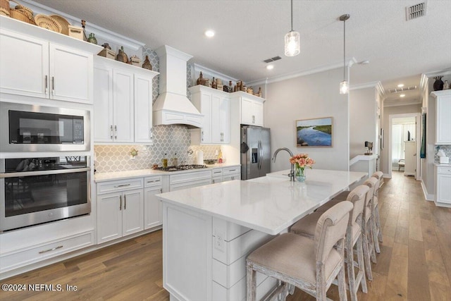 kitchen featuring premium range hood, a kitchen island with sink, hanging light fixtures, white cabinetry, and stainless steel appliances