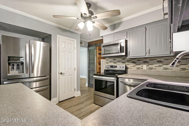 kitchen with a barn door, gray cabinetry, appliances with stainless steel finishes, decorative backsplash, and crown molding