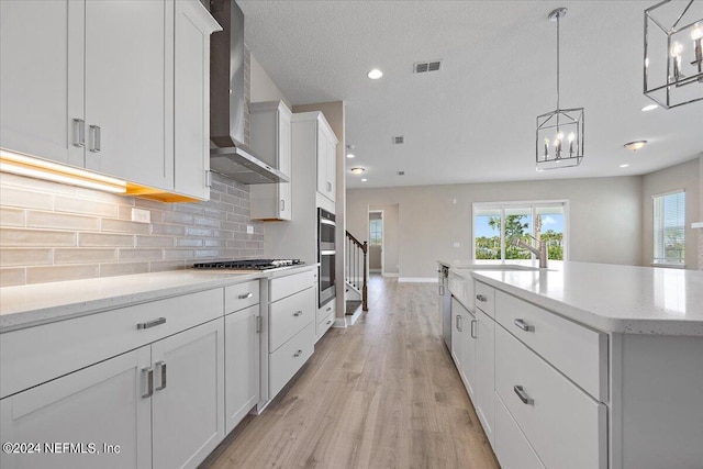 kitchen with a center island with sink, hanging light fixtures, white cabinets, wall chimney range hood, and backsplash