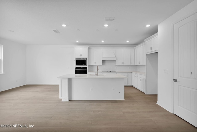 kitchen with light wood-type flooring, black appliances, a kitchen island with sink, a sink, and light countertops