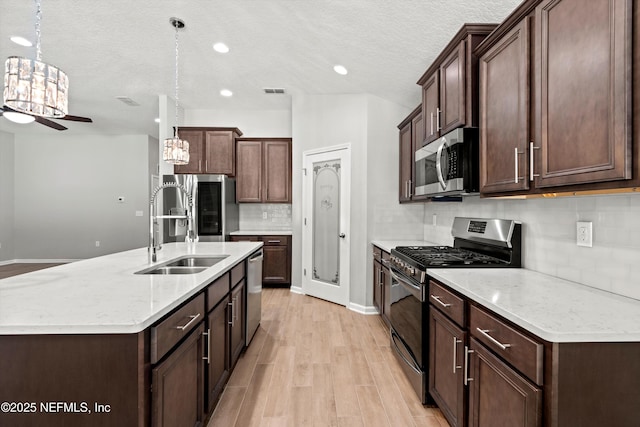 kitchen with an island with sink, sink, hanging light fixtures, stainless steel appliances, and light wood-type flooring