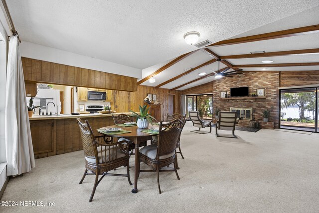 carpeted dining room featuring ceiling fan, a brick fireplace, vaulted ceiling with beams, wood walls, and a textured ceiling